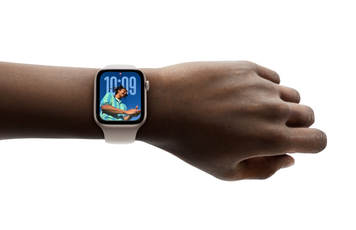 Primer plano de un Apple Watch en oro rosa con correa de goma en la muñeca de una persona que muestra la hora con el nuevo diseño en Liquid Glass