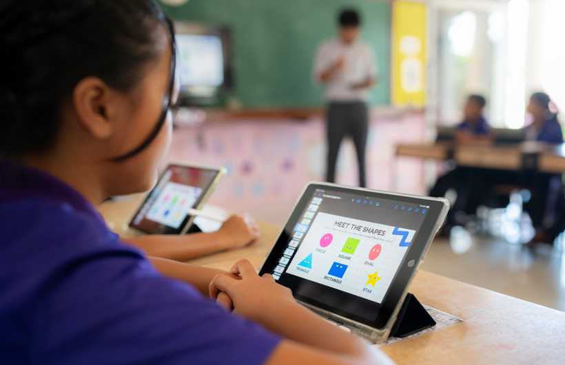 Primer plano de una estudiante en su sala con un iPad. En la pantalla hay unas figuras de colores con el título “Conoce las formas”.