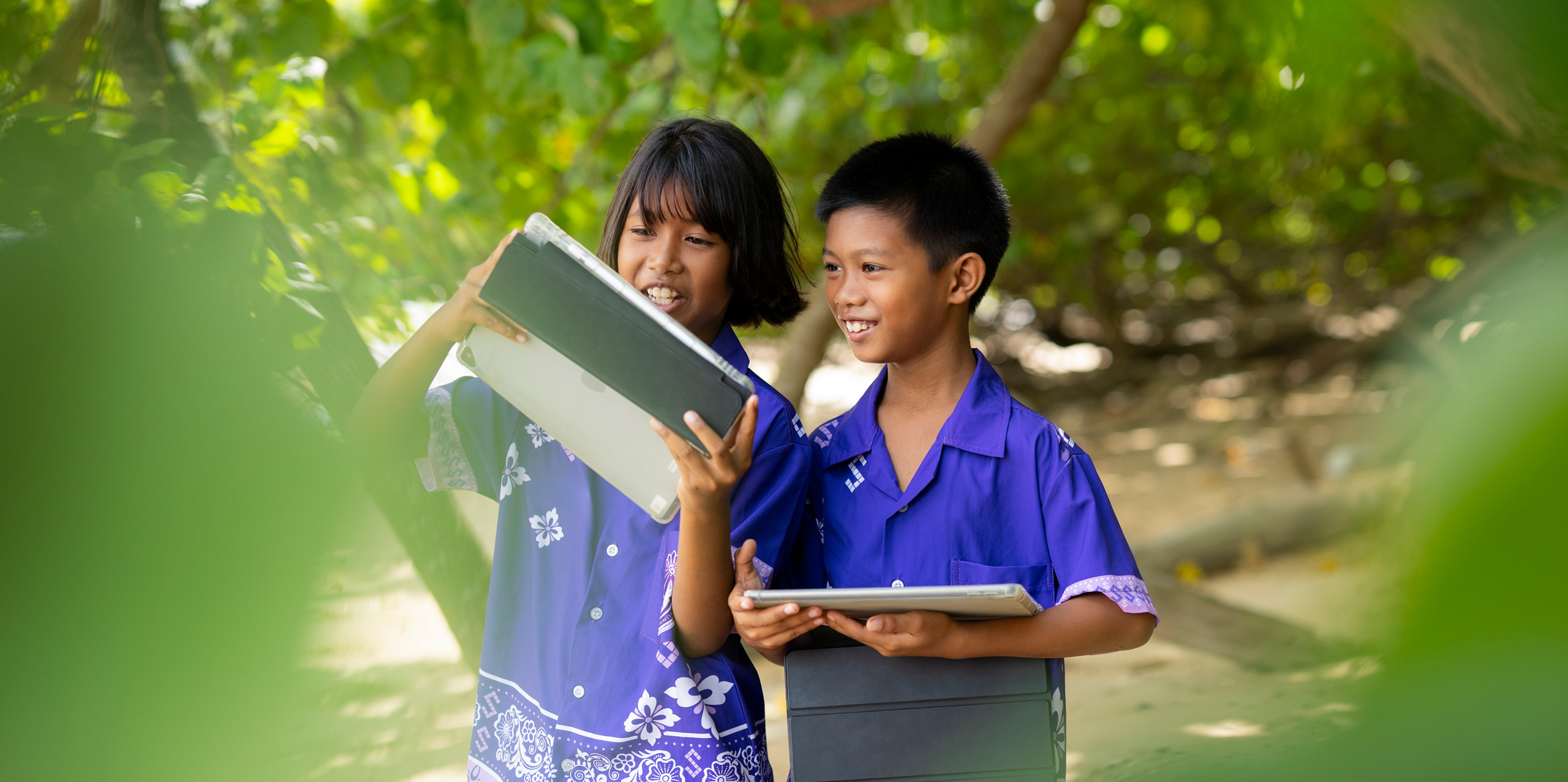 Dos estudiantes de primaria, un niño y una niña, de pie al aire libre entre árboles tropicales, cada uno con un iPad en la mano. Ambos sonríen y miran atentos la pantalla del iPad de la niña.